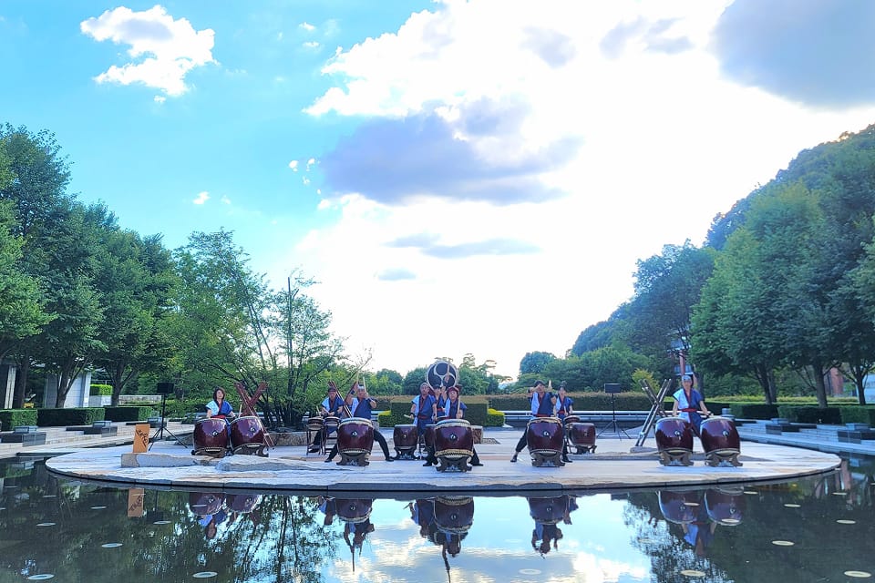 On a circular stage in the center of a water garden (reflecting pool), seven Japanese taiko drummers are performing. Taiko drums of various sizes are arranged across the stage, and performers dressed in traditional attire play with bachi (drumsticks). In the background, lush green trees, white clouds floating in a blue sky, and distant buildings are visible. The performing drummers, their drums, and the sky are all vividly reflected on the surface of the water in the foreground.