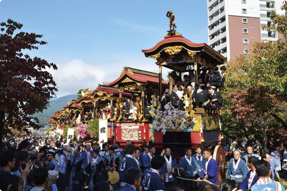 豪華な山車が並び、祭りの行列が街中を進む様子。多くの観客が集まり、にぎわいを見せている。