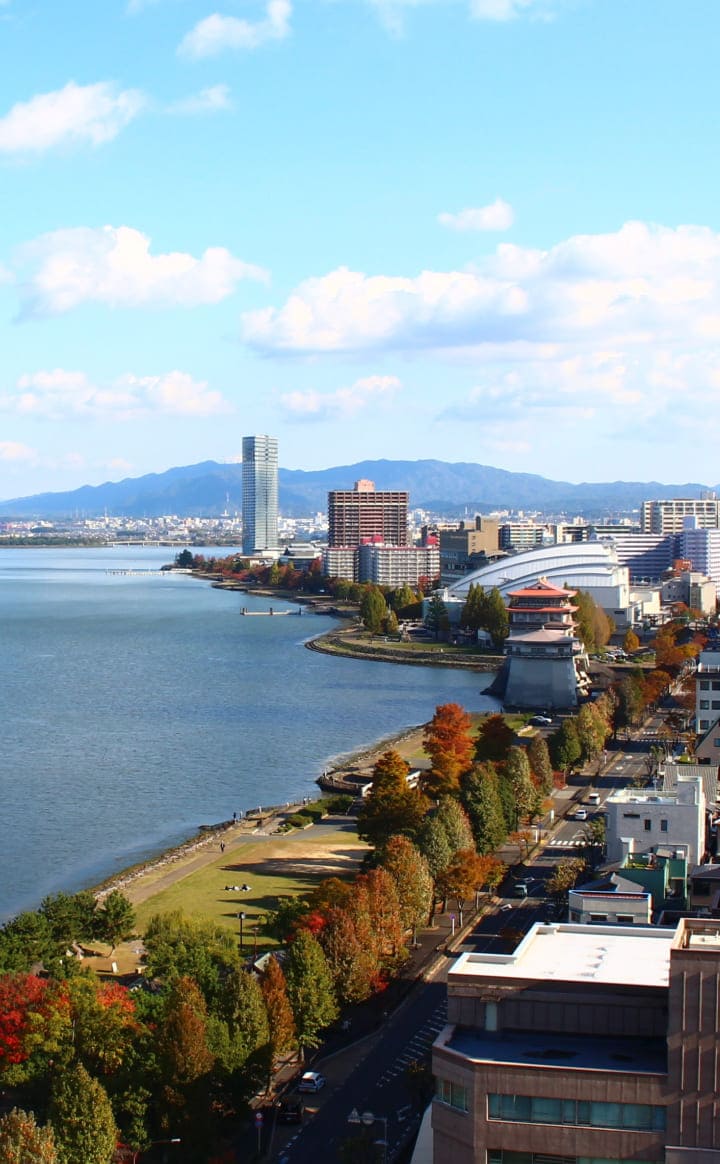 琵琶湖岸に広がる大津市の空撮写真。青く輝く琵琶湖と湖岸に立ち並ぶ近代的なビル群、背景には緑豊かな比叡山の山並みが広がっており、都市と自然が調和した風景です。