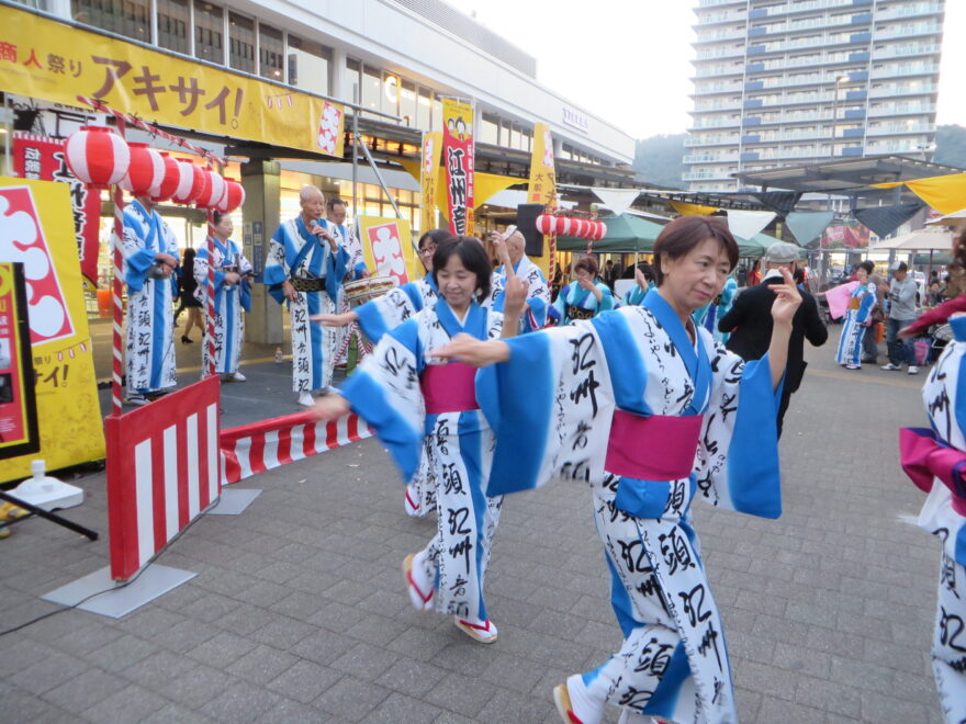 屋外で浴衣姿の女性たちが踊りを披露している様子。背景には祭りの装飾や屋台が並ぶ。